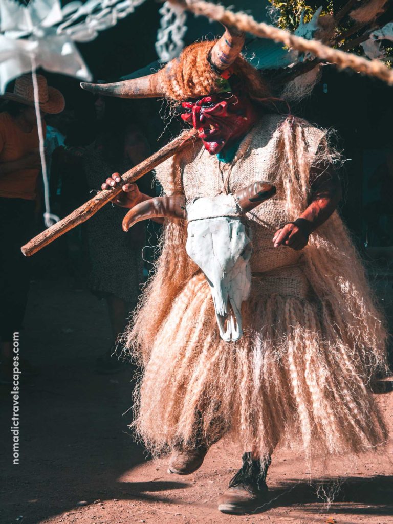 A Mexican Man Wearing A Red Demon Mask & Cow Skull, With Feathery White Dress
