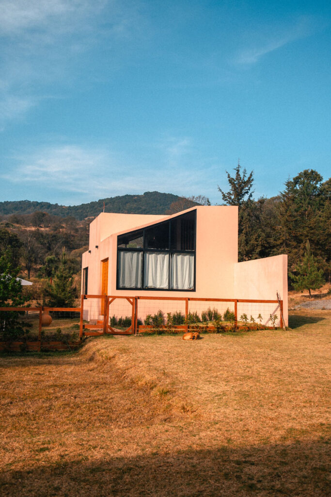 A pink Airbnb in the middle of the woods, with blue skies and yellow ground.