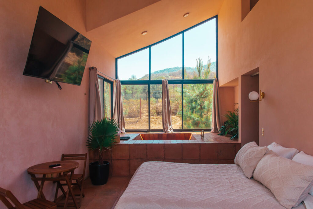 Pastel pink Airbnb of La Comuna, with a queen sided bed, tv on the left wall and a large window overlooking the forest
