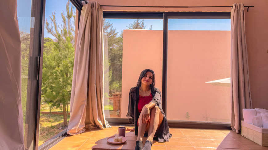 Travel Influencer Himanshi Shah wearing red PJS, sitting with her back to a large window with pink curtains